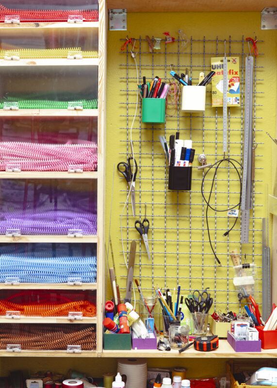 Drawers of colorful plastic spiral binding for books, next to a yellow wall with a metal grid holding different studio supplies.