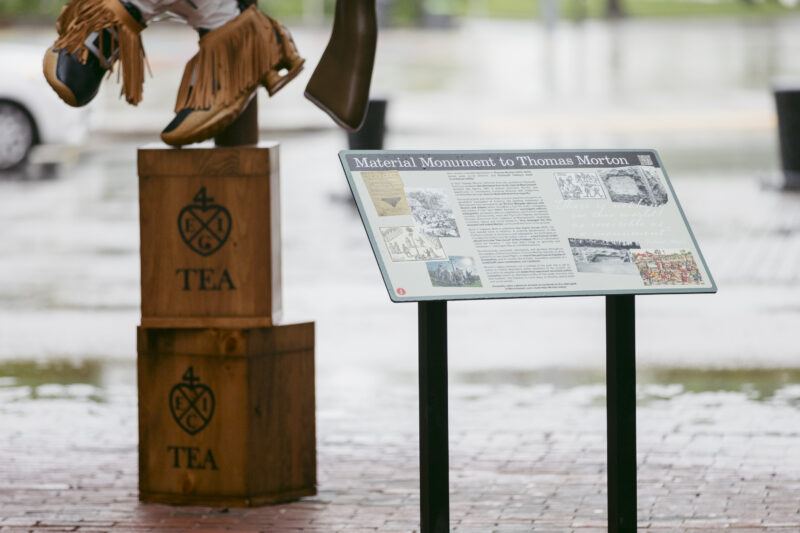 An informational sign titled "Material Monument to Thomas Morton" stands on brick pavement near two stacked wooden crates marked "TEA."