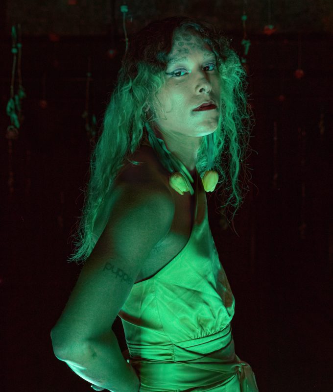 Jade Guanaro Kuriki-Olivo stands in profile, body turned to the right, head turned to face the camera, in a green light.