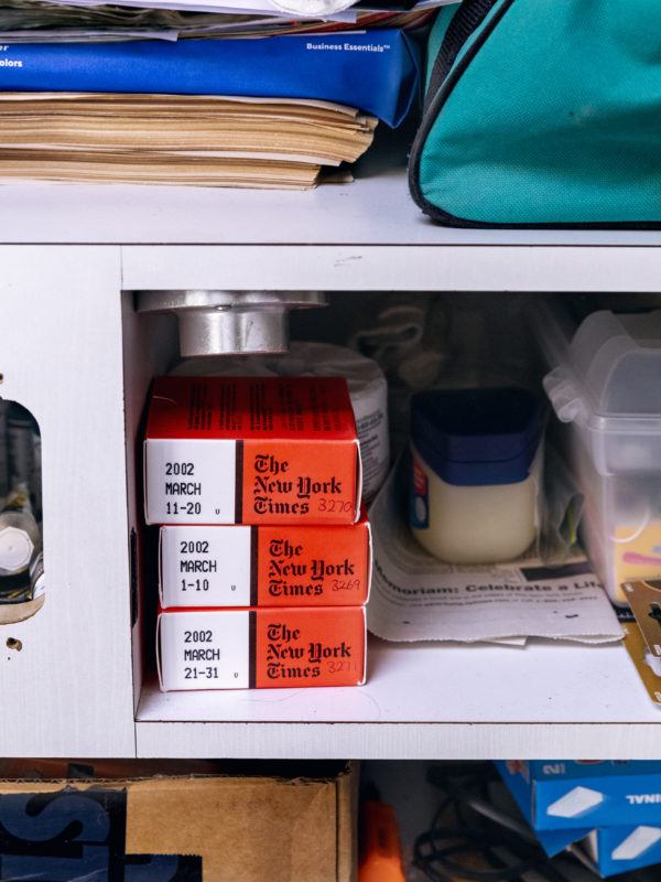 A shelf in Rose Salane's studio holds a variety of objects, the focus of the image is three small boxes which are predominately orange-red with one block of white, carrying microfilm of archival New York Times daily newspapers.