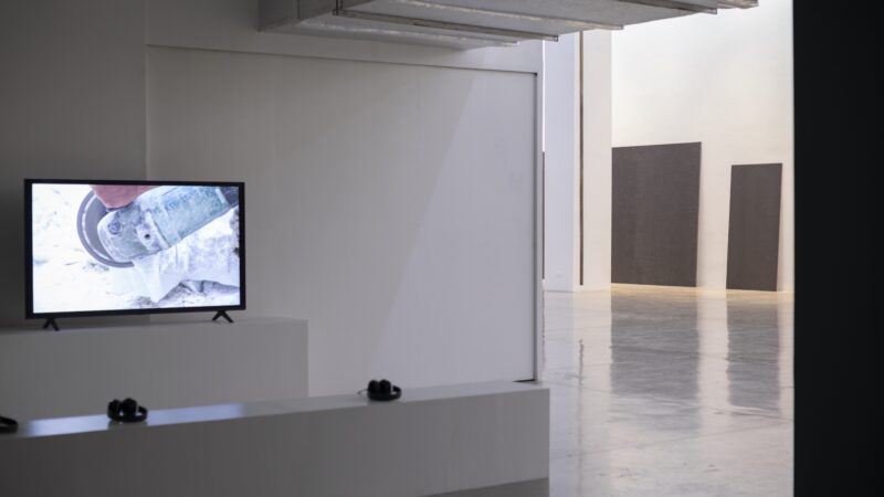 A wall-mounted television displays a close-up of a person using an angle grinder; in the gallery space behind two dark rectangular panels hang on a white wall.