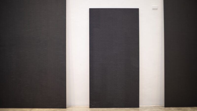 A large, rectangular, dark textured panel mounted vertically on a white gallery wall, flanked by two similar panels partially visible on either side.