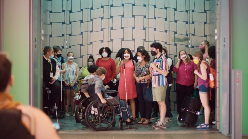 A diverse group of people, some wearing masks and one using a wheelchair, stand together in a large elevator with a geometric background.