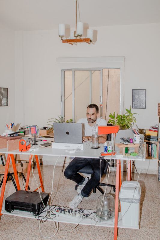 Artist Haig Aivazian sitting behind a desk looking at a computer, the desk is cluttered with objects like pens, paper, books, a lamp, and more, on the floor sits a printer, above Haig hangs a ceiling lamp that is off.