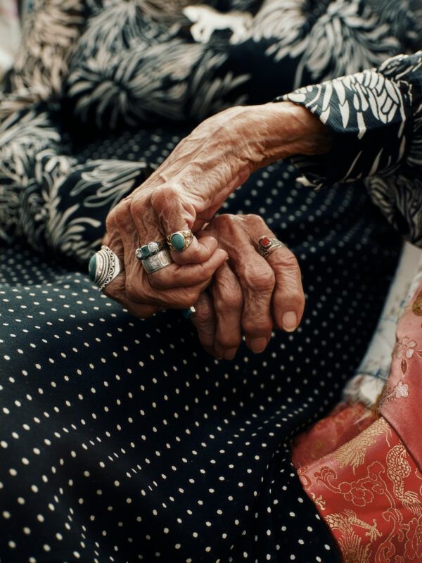 An older woman's hands resting in their lap, wearing rings and a patterned, polka-dotted dress with floral and embroidered fabric visible.
