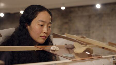 Hyeree Ro with long dark hair closely observes small sculpted objects displayed on a transparent surface supported by a wooden frame.
