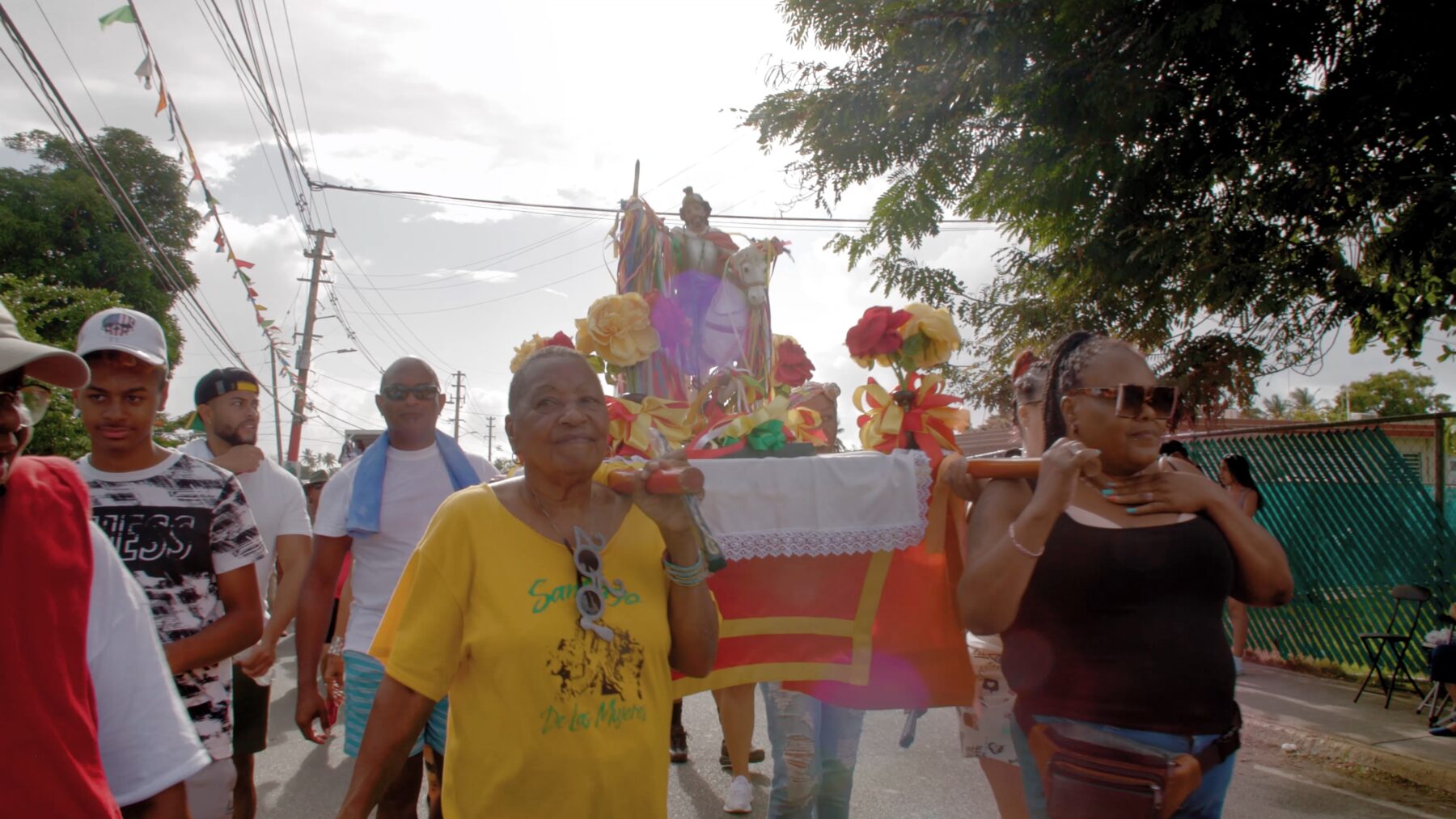 A group of people participate in a street procession, carrying a decorated float with flowers and religious figures under daylight.