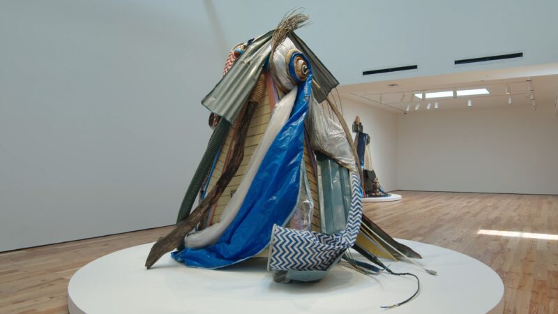 A large abstract sculpture composed of various colorful fabrics, tarps, and materials draped over a mound, displayed on a circular white platform in a gallery space.