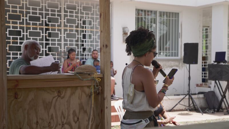 A woman speaks into a microphone while reading from her phone at an outdoor gathering; several people sit and listen in the background.