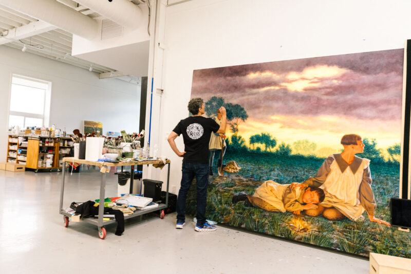Kent Monkman stands in a spacious studio, painting details on a large, realistic landscape artwork featuring three figures at sunset. Art supplies and storage shelves are visible in the background.