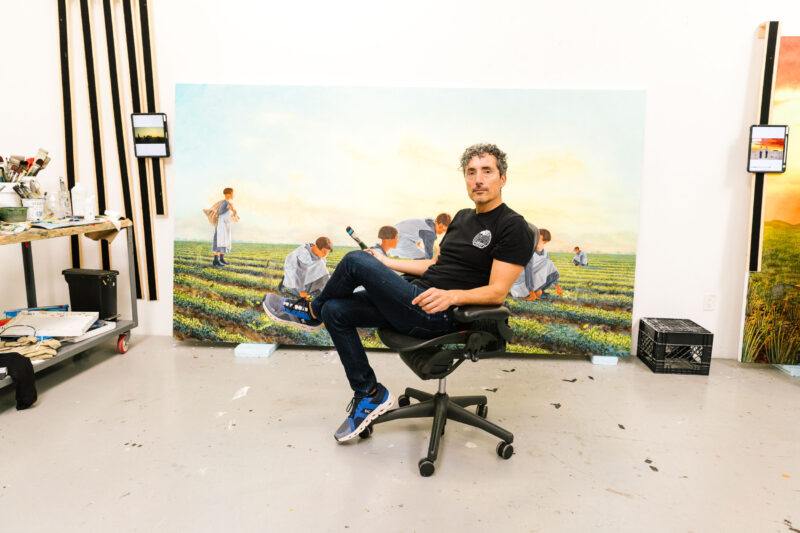 Kent Monkman sits on a chair in an art studio holding a paintbrush, with a large painting of people in a field displayed behind him and art supplies nearby.