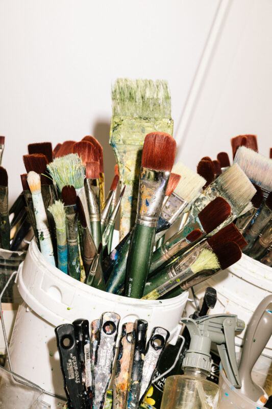A white container holds various paintbrushes with colorful bristles, surrounded by palette knives and a spray bottle on a table.