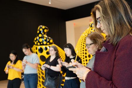 Six people stand in front of yellow, polka-dotted sculptures, focusing on writing or drawing on notepads or phones.