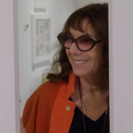 Sophie Calle with wavy brown hair wearing sunglasses and an orange top smiles while standing partially behind a white wall in an art gallery.