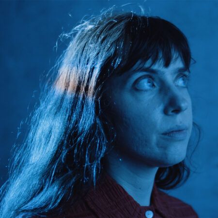 Jacky Connolly with long dark hair and bangs looks upward, illuminated by blue light, wearing a red button-up shirt against a blue background.