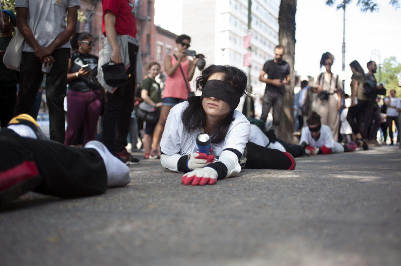 Participants crawling on the pavement as part of Pope.L’s "Conquest," they wear blindfolds and gloves, and are holding flashlights.