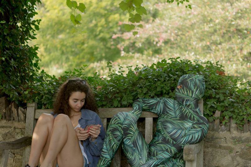 A woman sits on a bench looking at her phone, next to a person in a leaf-patterned bodysuit. Lush greenery is visible in the background.