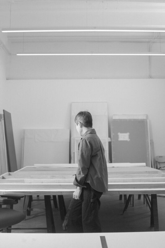 Black and white image of Maria Taniguchi standing next to a large table in a spacious, well-lit room with various boards leaning against the wall.