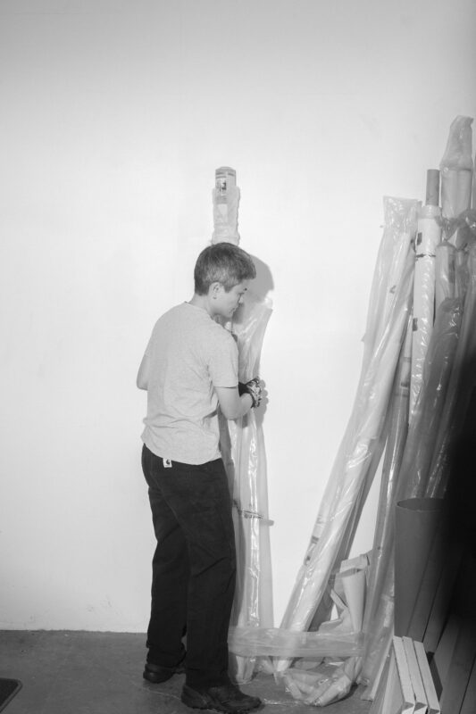 Black and White image of Maria Taniguchi holding a long piece of material wrapped in plastic leaning against a wall.