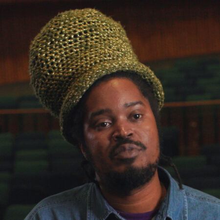 Julien Creuzet with a beard and a tall crocheted hat sits in an empty auditorium.