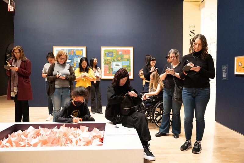 A group of people in an art gallery stand and sit near an exhibit, writing on notepads. Artworks are displayed on the walls in the background.