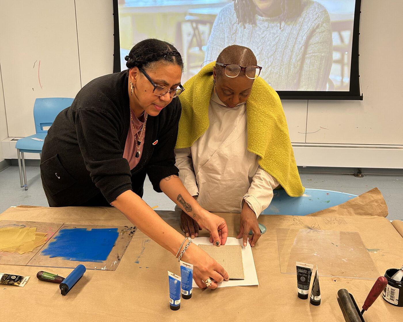 Two people stand at a table working together on an art project with paint tubes and paper, while a projected image is visible in the background.