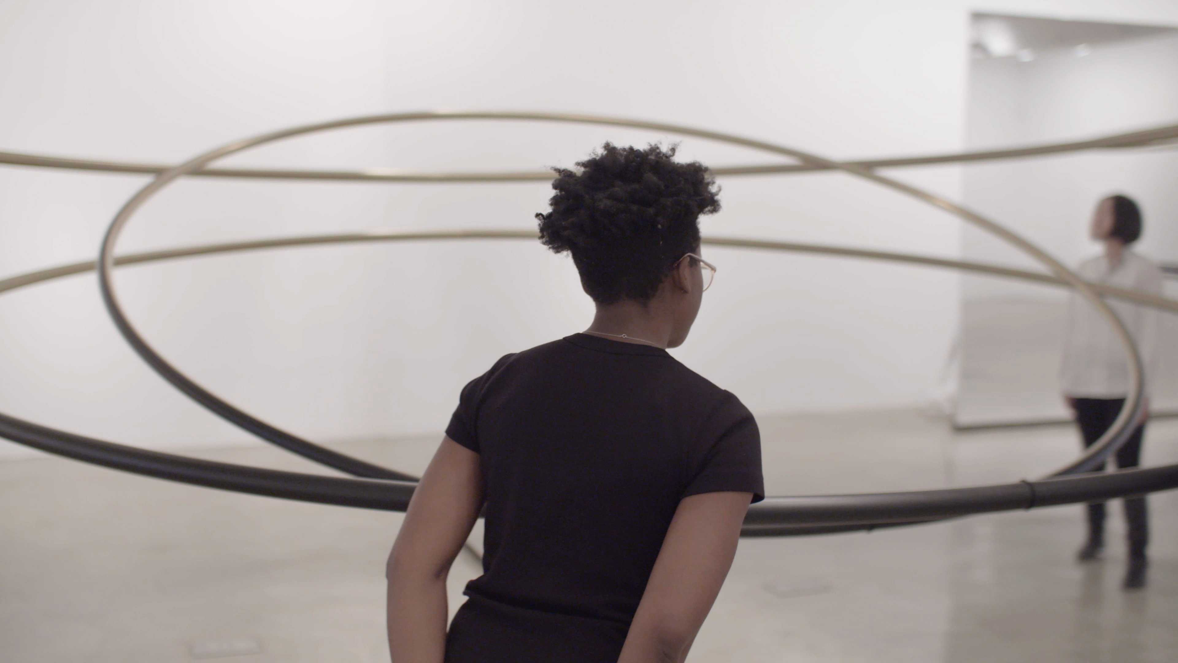 A person stands in front of a large, circular metal sculpture in a modern art gallery; another person is seen in the background.