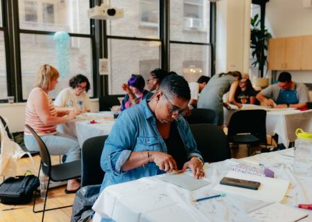 People seated at tables in a well-lit room work on arts and crafts projects, using tools and materials spread out in front of them.