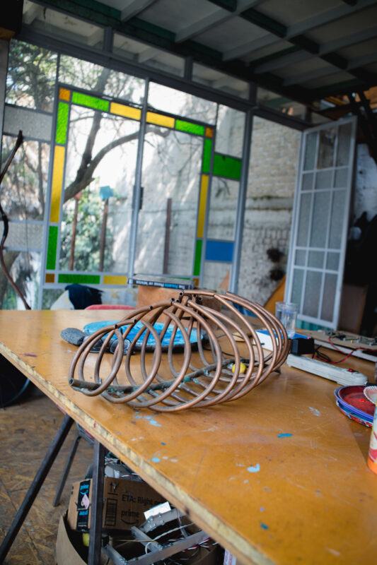 A metal sculpture on a wooden table in a sunlit room with stained glass windows and various tools scattered around.