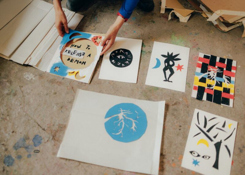 Cut-out drawings laid on the floor of the studio, Tamar Ettun's hands are mid placing one of them down on the ground.