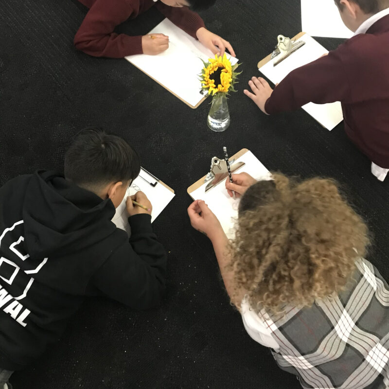 Four children lying on the floor, drawing on clipboards, with a vase of yellow flowers placed nearby on a dark carpet.