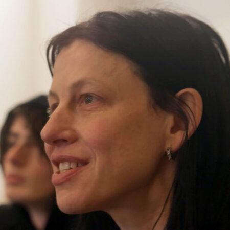 Anne Imhof smiling slightly and looking forward, with a figure out of focus behind her.