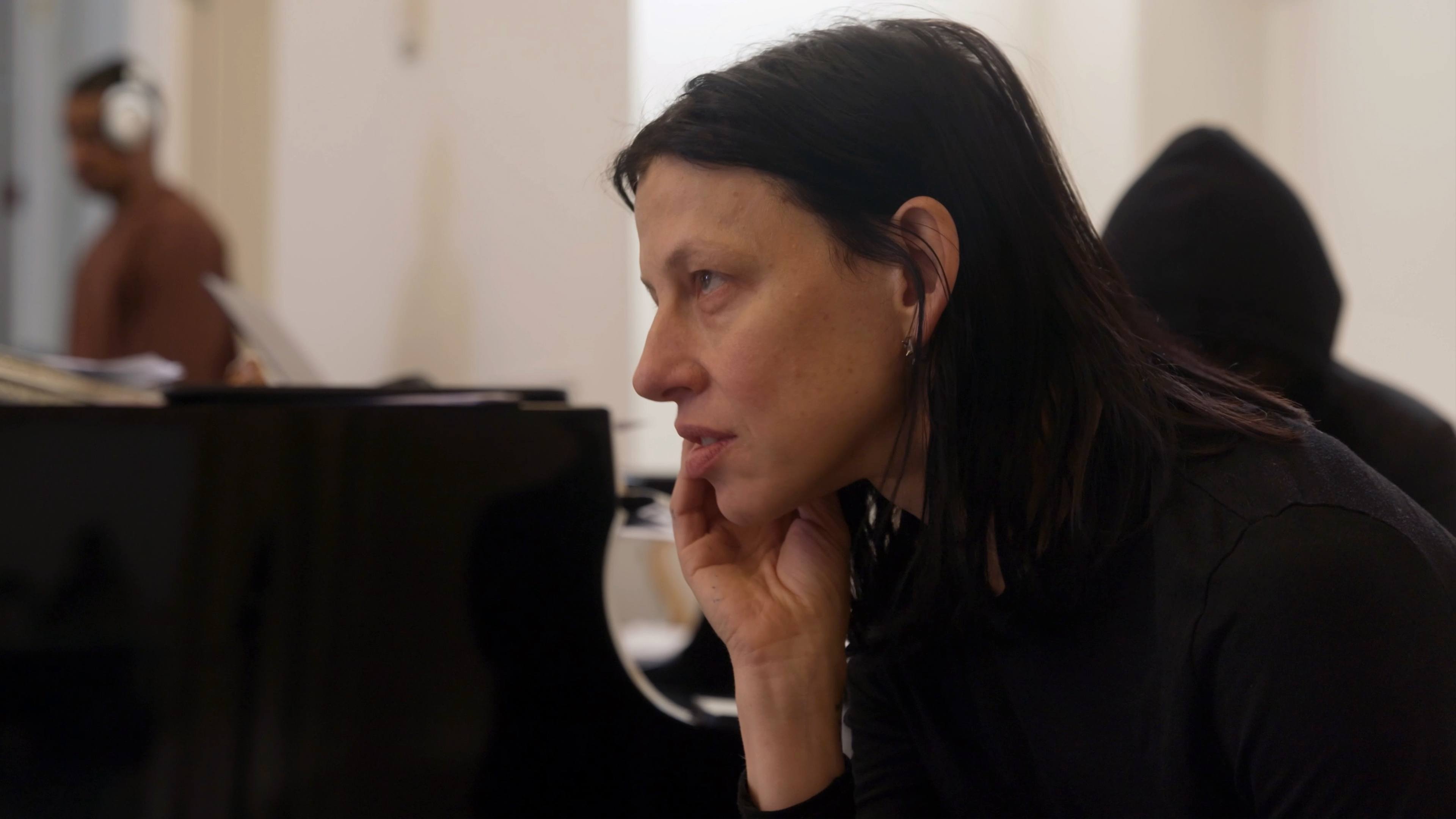 Anne Imhof holding her head in her hand looking forward, with a piano next to her.