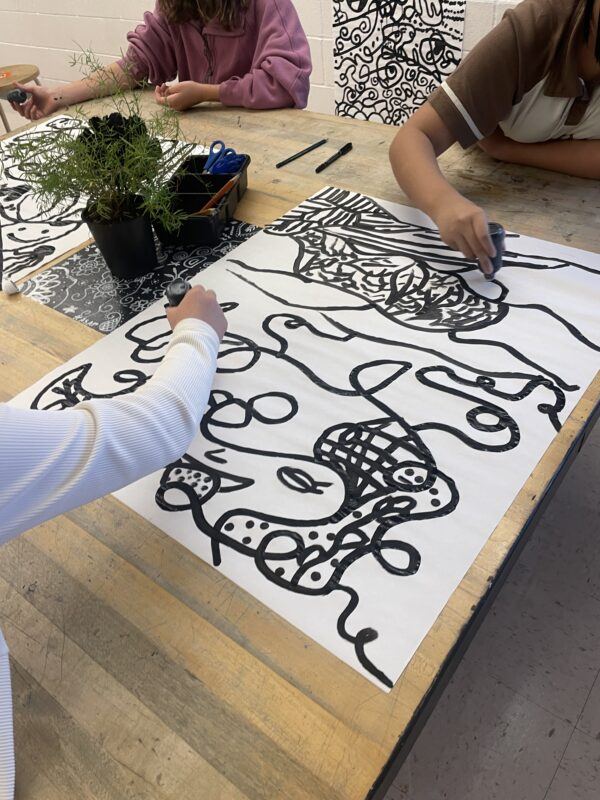 Three children seated at a table use black markers to draw abstract line designs on large sheets of white paper; a small potted plant and art supplies are on the table.