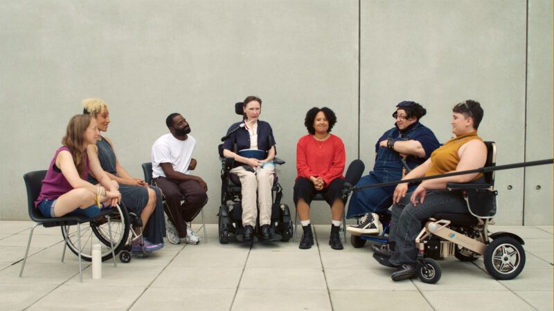 A diverse group of seven people, some using wheelchairs, sit in a semicircle outside against a plain concrete wall, engaged in conversation.