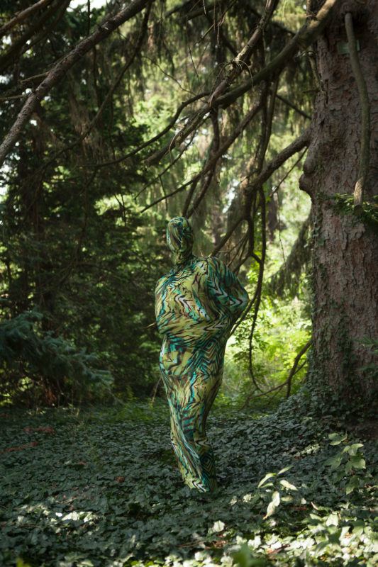 A person camouflaged in a green and brown patterned suit stands in a dense, leafy forest with large trees and hanging branches.