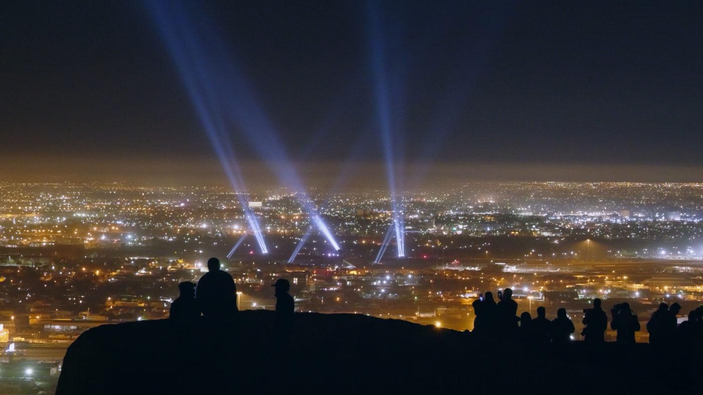 Silhouetted people watch city lights and spotlights in the night sky from a hilltop.