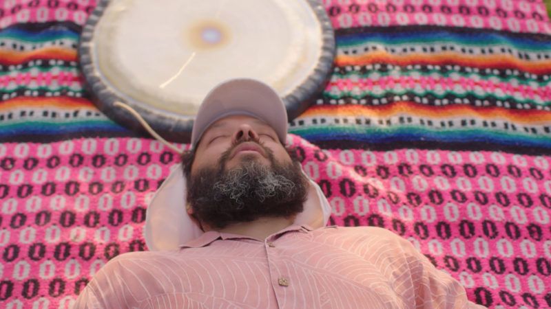 Guadalupe Maravilla wearing a pink button-up shirt and white hat, laying down on a colorful blanket with his eyes closed. 