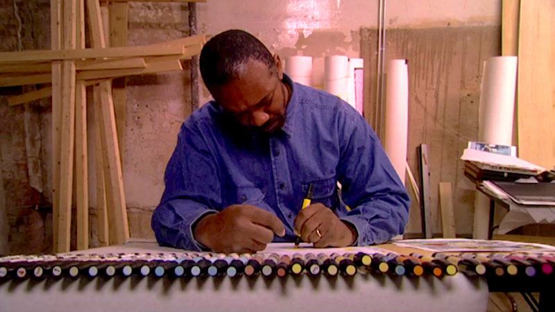 Kerry James Marshall in his studio wearing a denim button up shirt, drawing with a yellow marker on a white sheet of paper. A neat line of markers sit in front of him, balanced on the edge of the table.