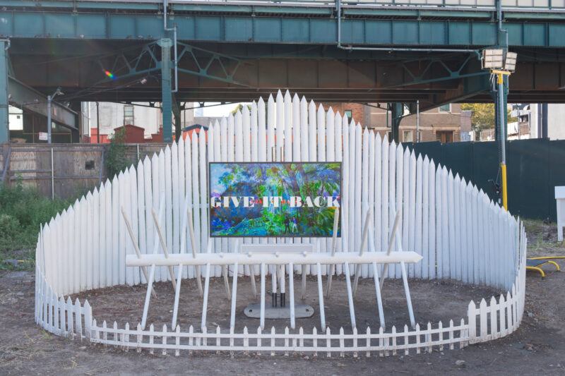 A digital screen displaying "GIVE IT BACK" is surrounded by a pointed white fence in an outdoor urban area, with a raised train track in the background.