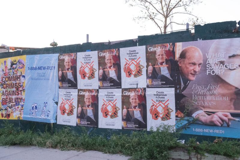 A row of posters on a fence reading "Give it back" posters featuring a man giving thumbs up, with text referencing Indigenous land.