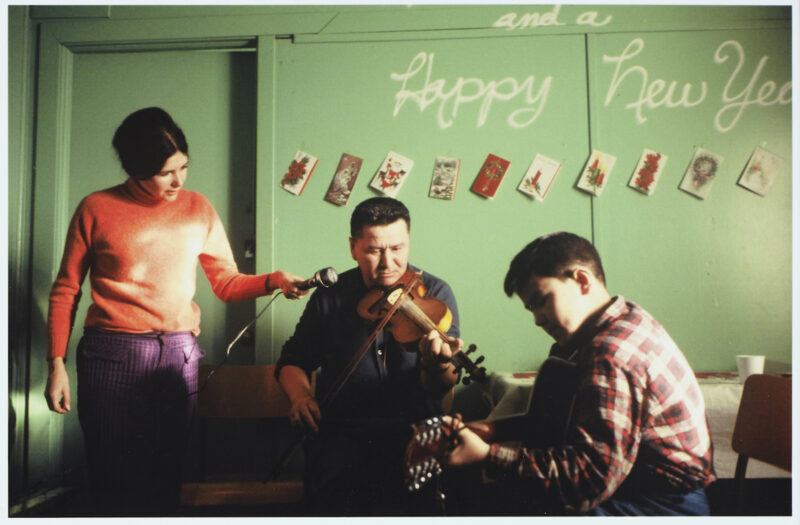 A woman holds a microphone for a man playing the violin while another man plays guitar. Holiday cards and "Happy New Year" are displayed on a green wall behind them.