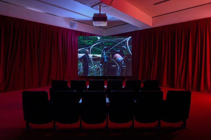 A small theater room with red curtains and empty black chairs faces a large screen displaying an image of barbed wire and greenery.