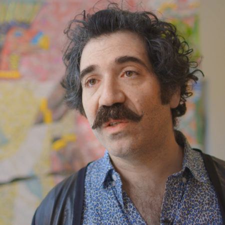Michael Rakowitz with a mustache and curly hair, wearing a blue patterned shirt and jacket.