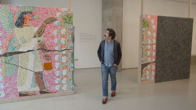Michael Rakowitz observes his vibrant, large-scale artworks displayed in a gallery, with one featuring a colorful, mythical figure on a floral background.