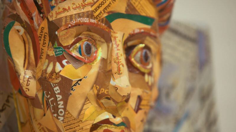 A close-up of a sculpture made from recycled paper and cardboard, featuring colorful fragments of text and imagery on a human head form.