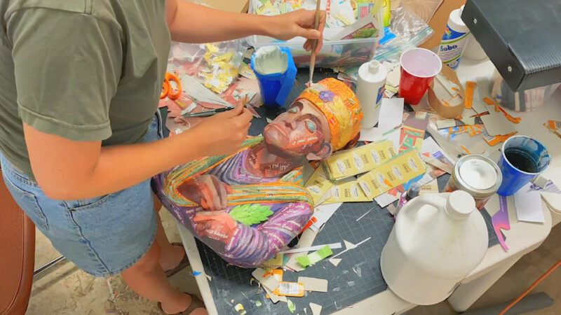 Person painting a colorful bust on a cluttered table with brushes, paints, and crafting materials scattered around.
