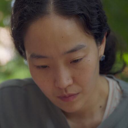 Hyeree Ro with dark hair looks down with a neutral expression, in an outdoor setting.