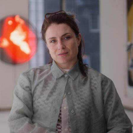 Rachel Rossin with brown hair, wearing a sheer gray button-up shirt, sits indoors with blurred abstract art in the background.