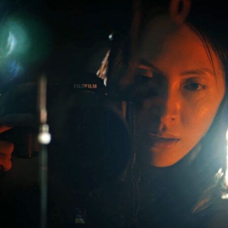 Diane Severin Nguyen holding a Fujifilm camera, partially illuminated with warm light, looking directly into the lens in a dimly lit setting.